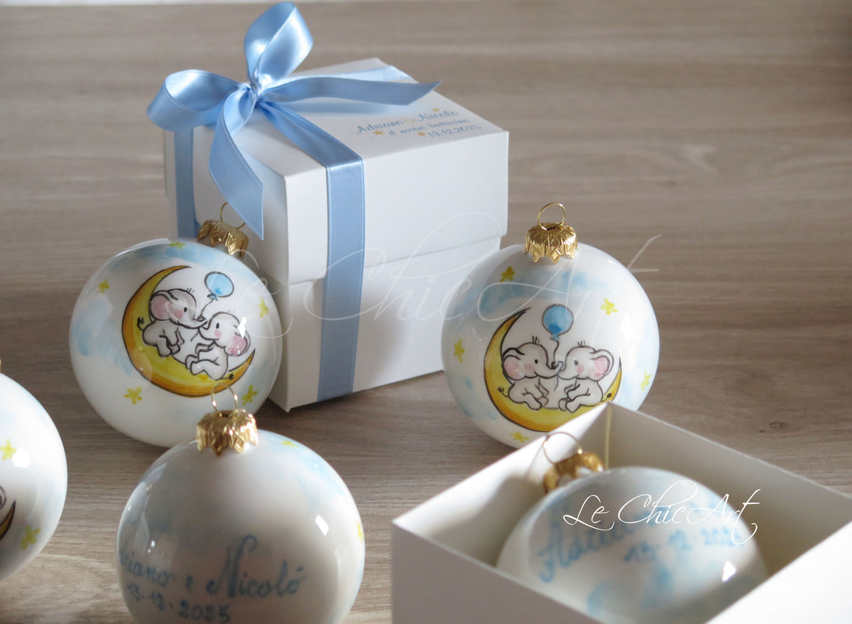 Palle in ceramica per Battesimo di gemelli, dipinte a mano con due elefantini sulla luna gialla, palloncino azzurro e stelline, accanto alla scatola con fiocco azzurro.
