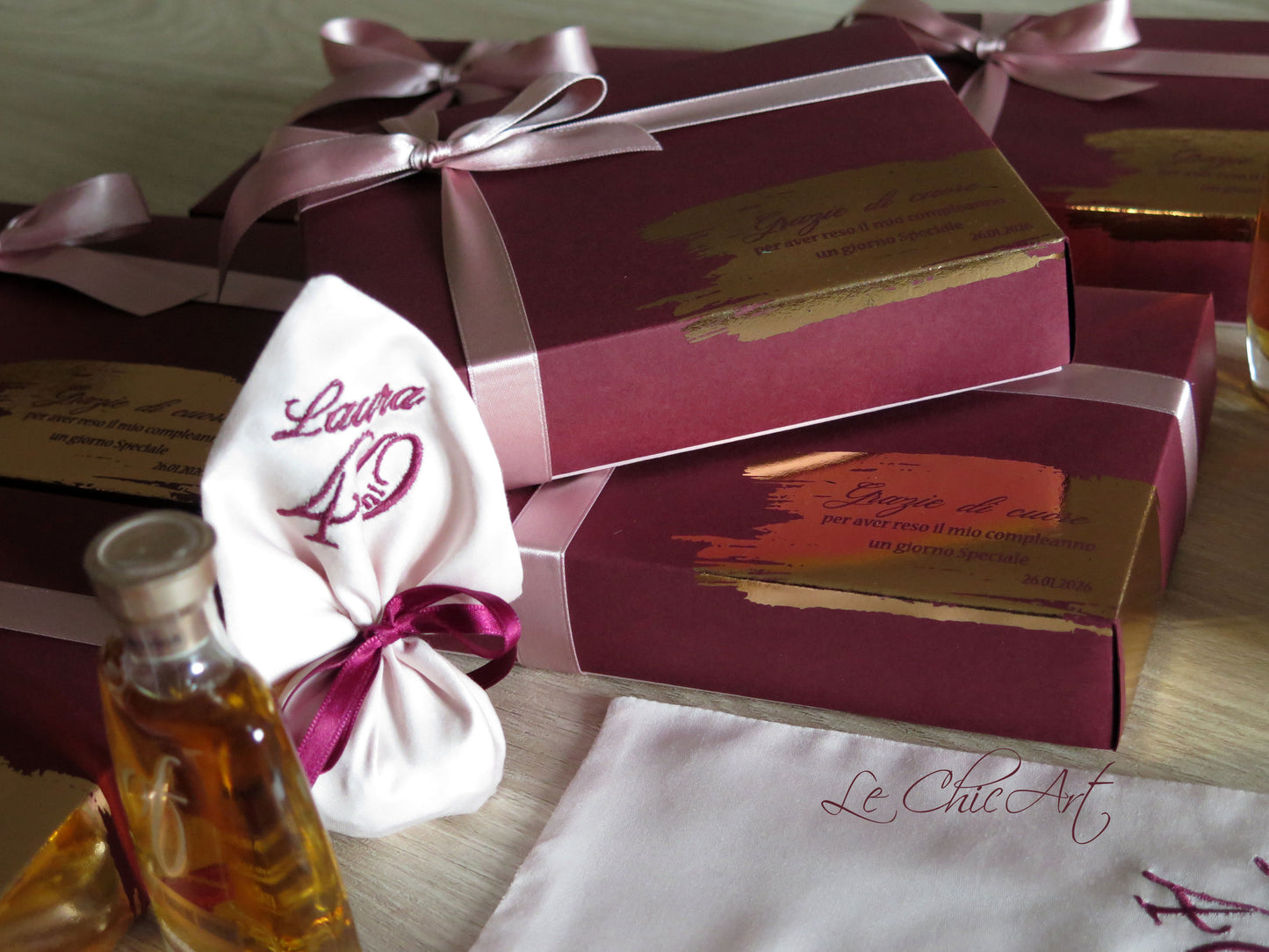 Composizione ravvicinata di diverse scatole regalo borgogna con fiocchi rosa antico e inserti in lamina oro rosa. In primo piano, un sacchetto di raso rosa cipria ricamato con 'Laura 40' e una mini bottiglia di grappa sfocata