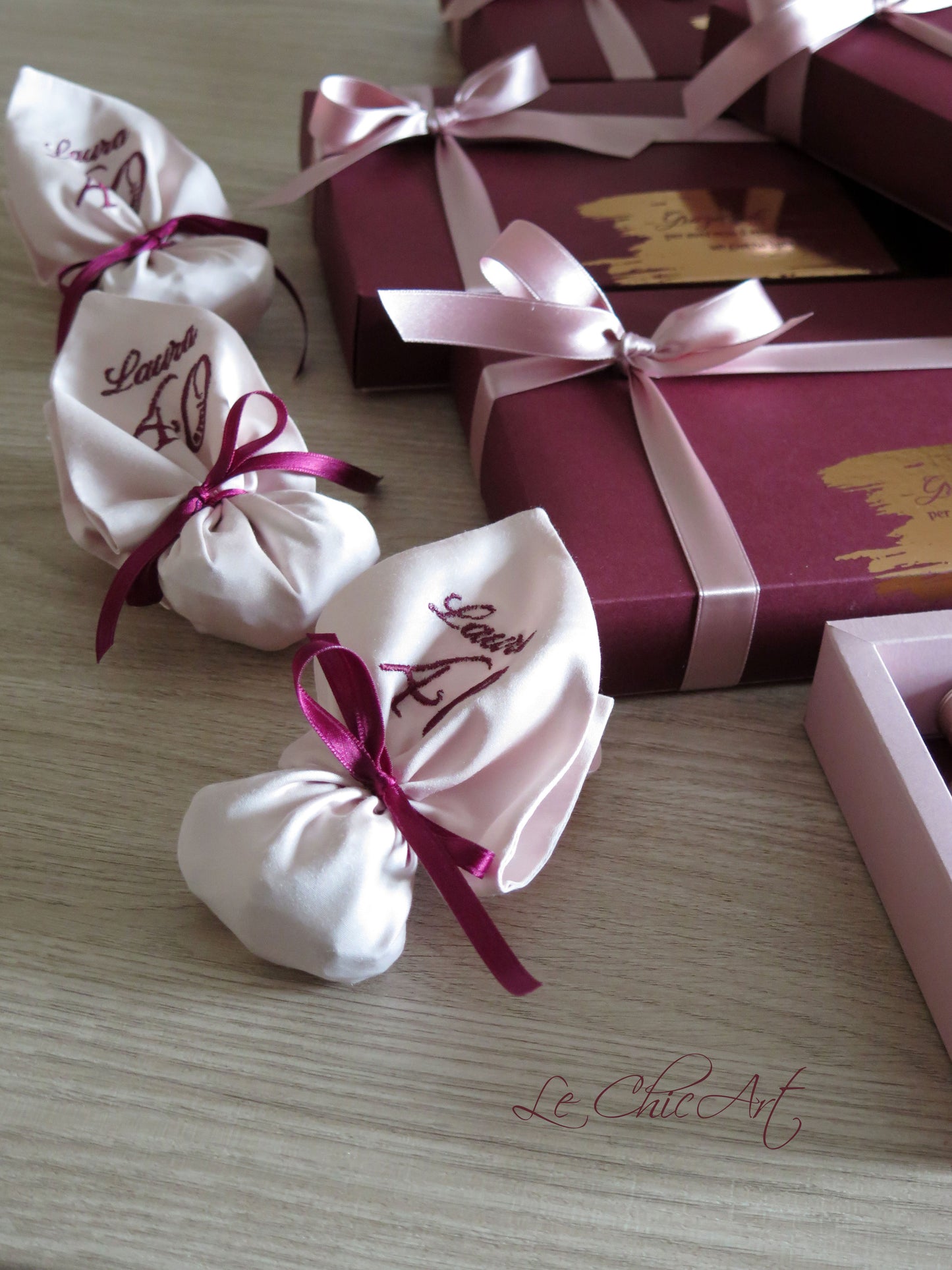Primo piano di tre sacchetti di raso rosa cipria, ricamati con 'Laura 40' e legati con nastri color borgogna, appoggiati su un tavolo di legno chiaro, con bomboniere borgogna sullo sfondo