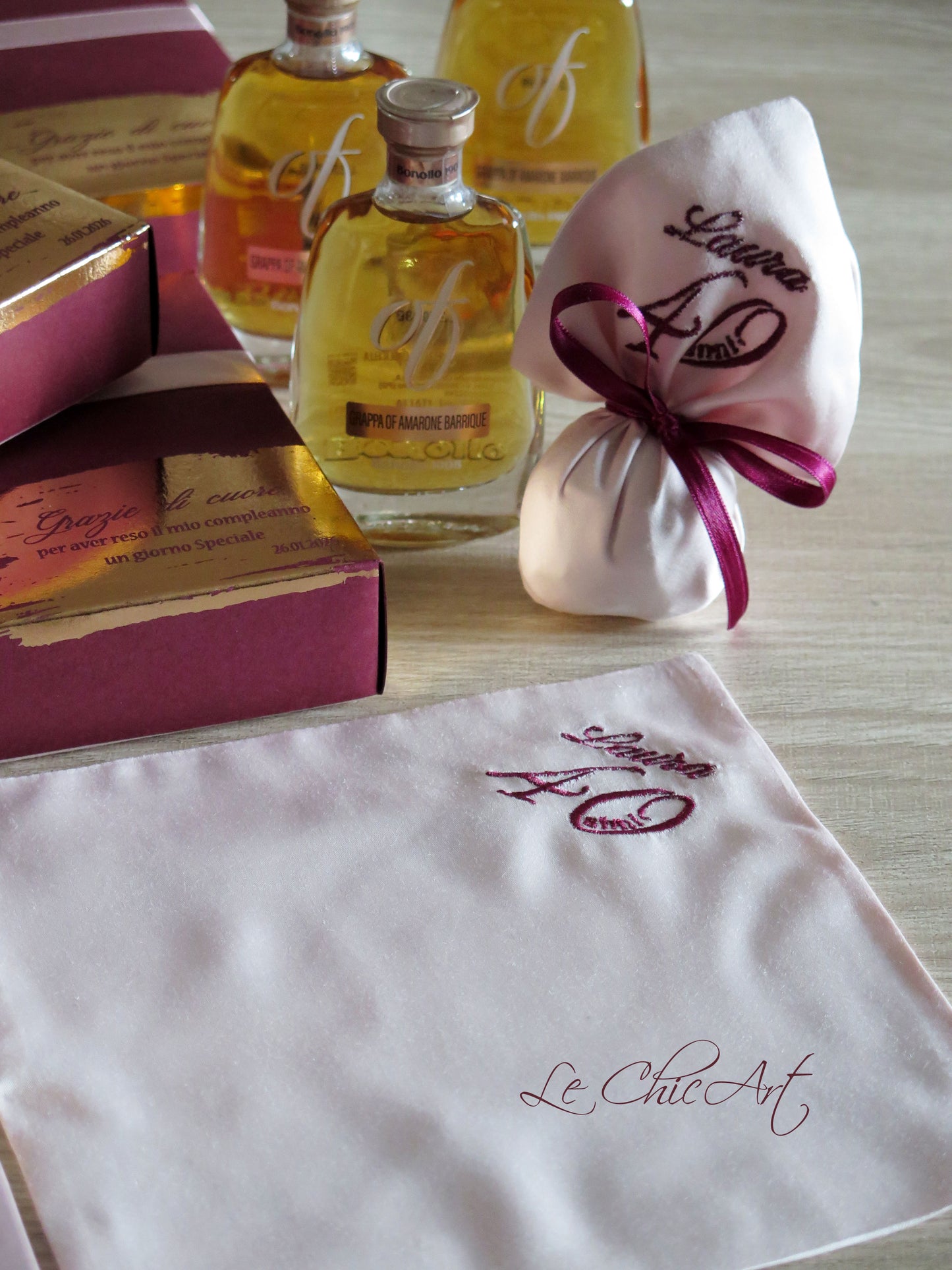 Dettaglio di un sacchetto di raso rosa cipria ricamato con 'Laura 40' e bottiglie di grappa OF Bonollo sullo sfondo. In primo piano, un pezzo di tessuto di raso con il logo 'Le Chic Art'