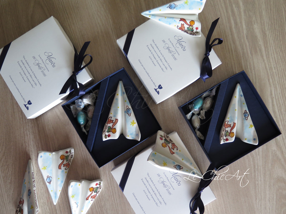 Vista dall’alto delle bomboniere aeroplano in ceramica con Piccolo Principe, confezioni blu notte e coperchi bianchi con stampa per la Prima Comunione.