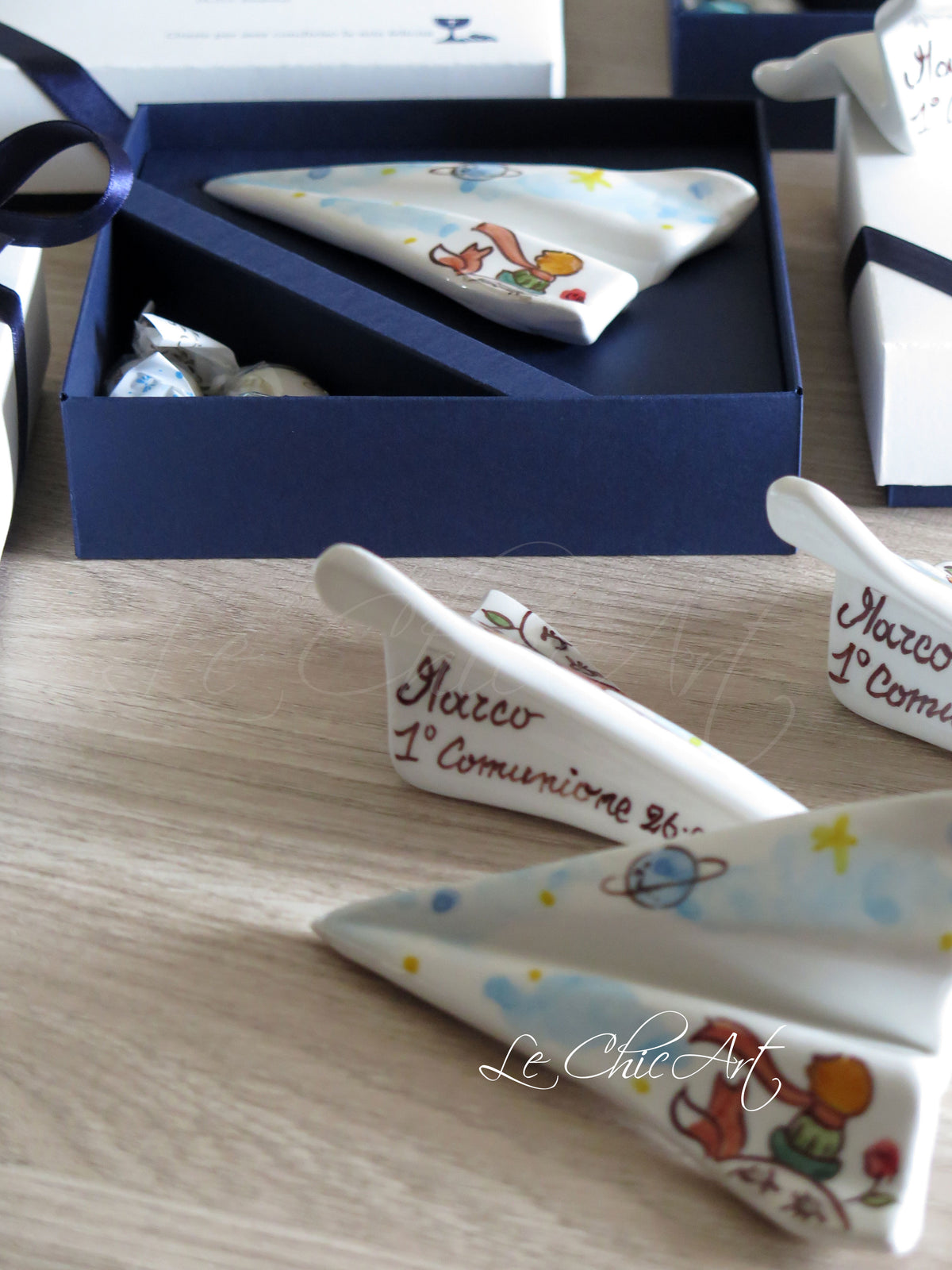 Aeroplano in ceramica dipinto a mano con Piccolo Principe, volpe e pianeti, con nome e scritta 1° Comunione sotto l’ala, accanto alla scatola blu notte.
