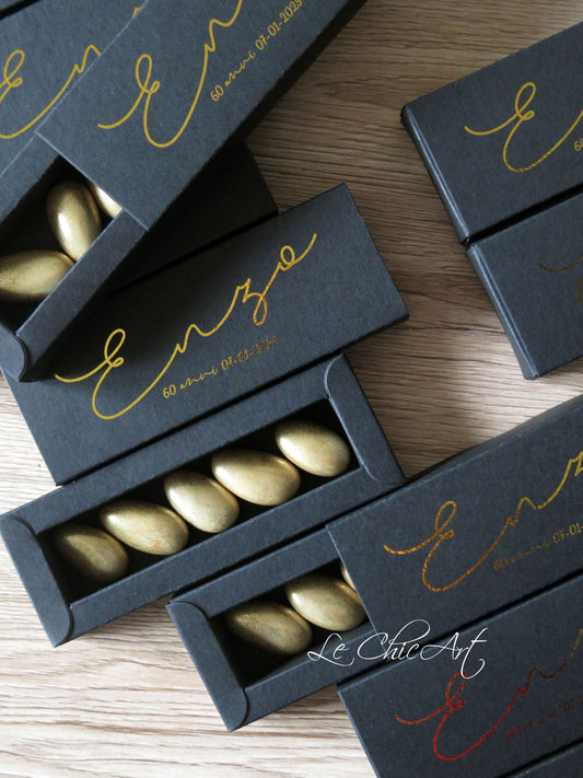 Set di scatole porta confetti nere con stampa oro lucido per 60° compleanno, disposte su superficie in legno, sistema a sfilo elegante