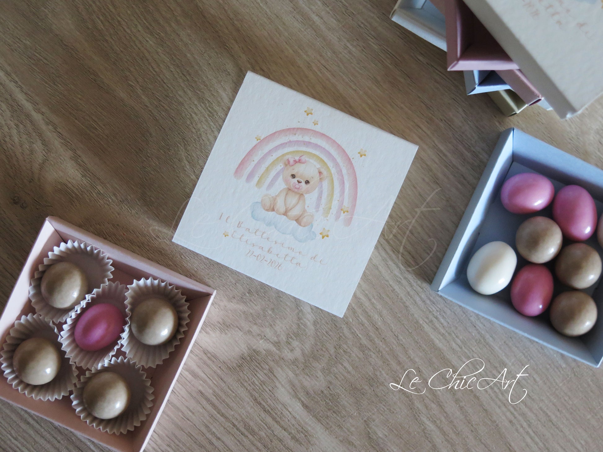 Dettaglio scatolina degustazione confetti con illustrazione orsetta femmina seduta su arcobaleno con stelline dorate e nuvole soffici, fondo rosa cipria, struttura a cassetto con vassoio interno colorato 8x8x2 cm